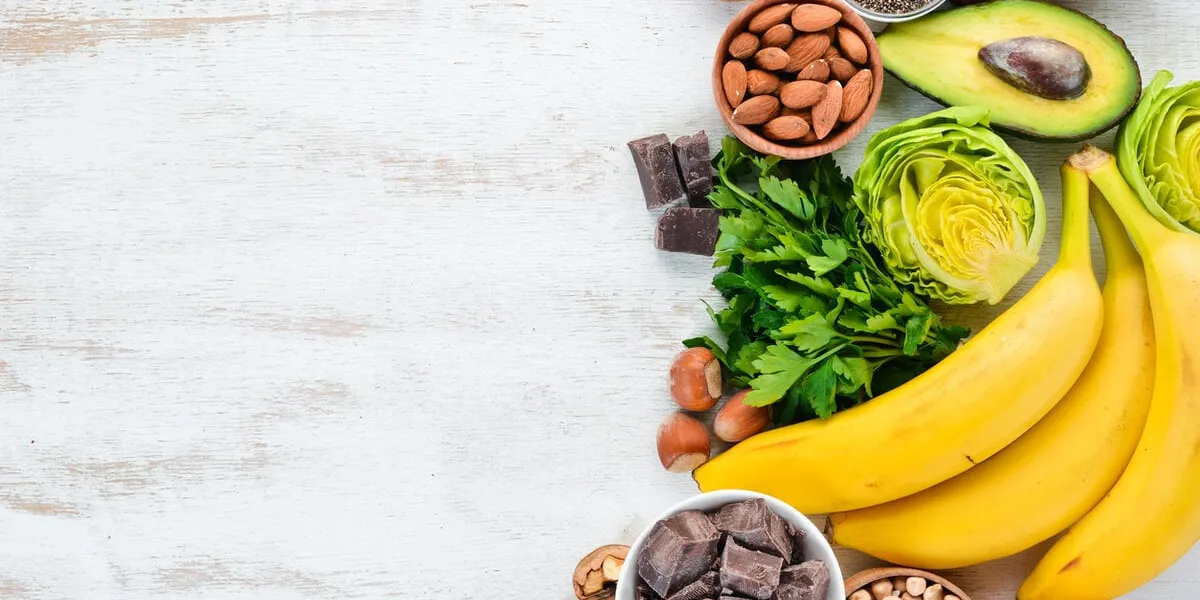 foods containing natural magnesium mg  chocolate, banana, cocoa, nuts, avocados, broccoli, almonds top view on a white wooden background