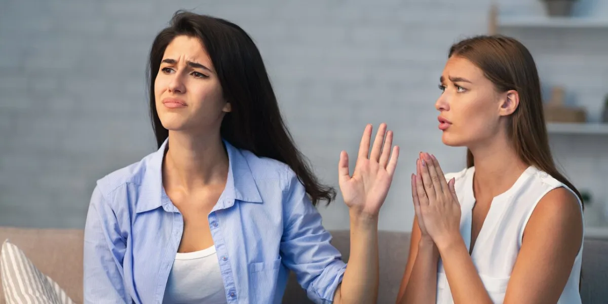 miserable girl asking indifferent friend for a favor begging holding hands in prayer gesture sitting on sofa indoor selective focus