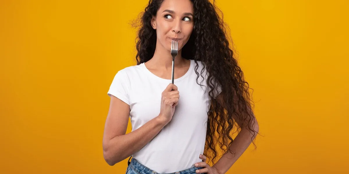 portrait of excited hungry young woman holding fork in her mouth, biting and dreaming about delicious healthy food, choosing something to eat posing looking aside isolated on yellow orange studio wall