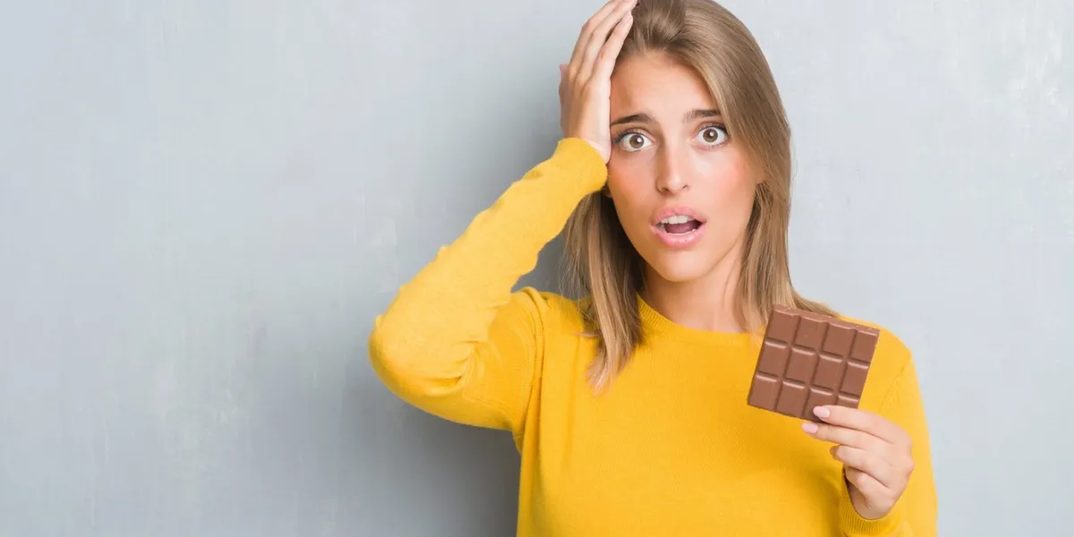 beautiful young woman over grunge grey wall eating chocolate bar stressed with hand on head, shocked with shame and surprise face, angry and frustrated fear and upset for mistake