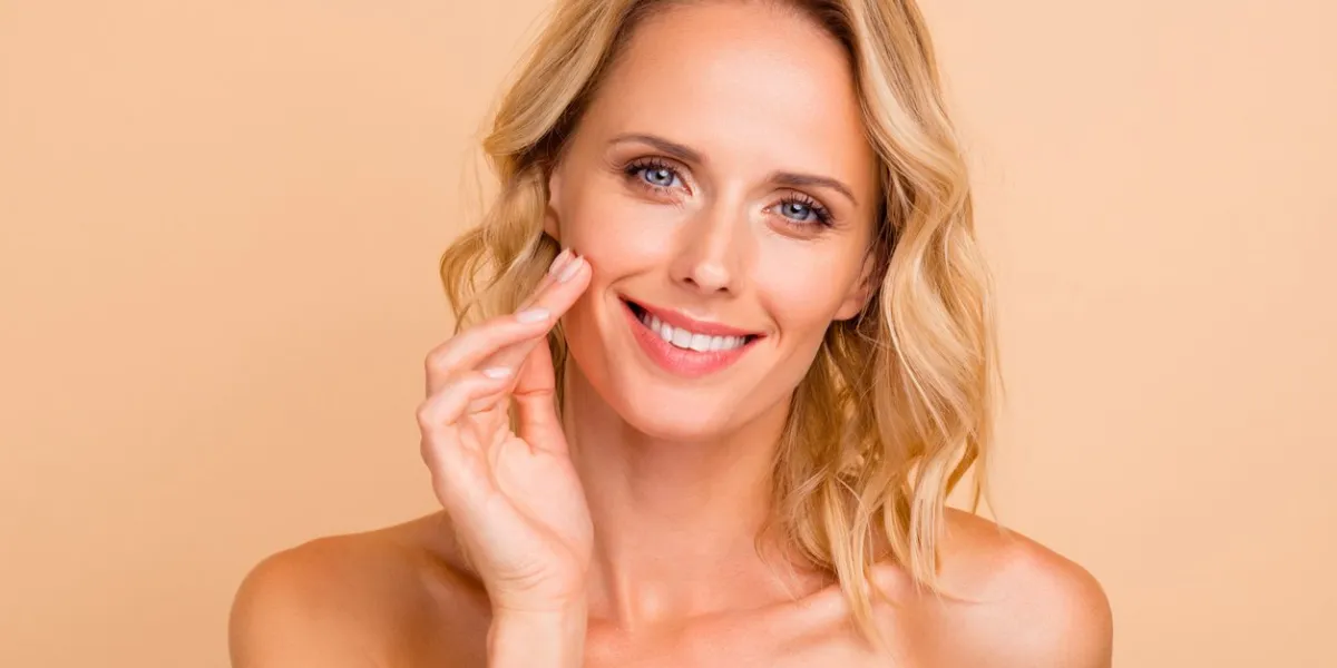 salon spa therapy treatment concept close-up portrait of attractive cheerful wavy-haired lady with flawless smooth pure clean clear skin applying cream on cheek isolated on beige background