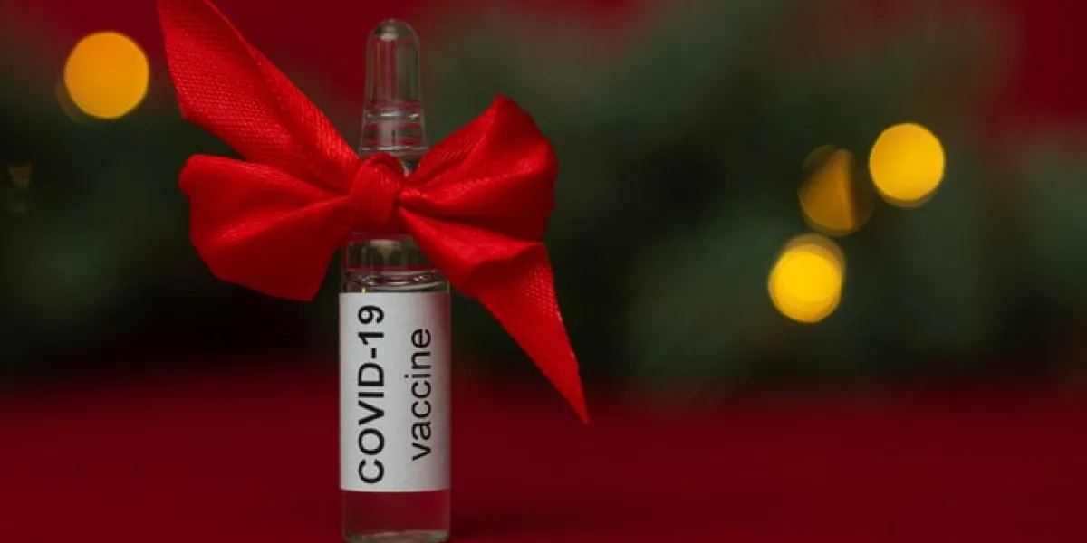 an ampoule of the covid-19 coronavirus vaccine stands as a gift near a christmas tree branch with lights on a red background