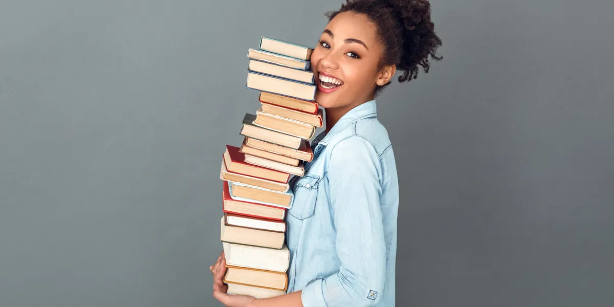 young african female student isolated on grey wall holding a pile of books standing profile looking camera laughing