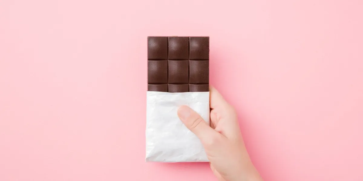 young woman hand holding chocolate bar on light pink table background opened pack sweet snack closeup top down view