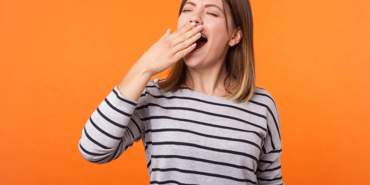 portrait of exhausted or bored young woman with brown hair in long sleeve striped shirt standing, yawning and covering mouth with hand, disinterest indoor studio shot isolated on orange background