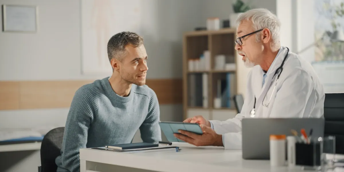 experienced middle aged family doctor showing analysis results on tablet computer to male patient during consultation in a health clinic physician sitting behind a desk in hospital office