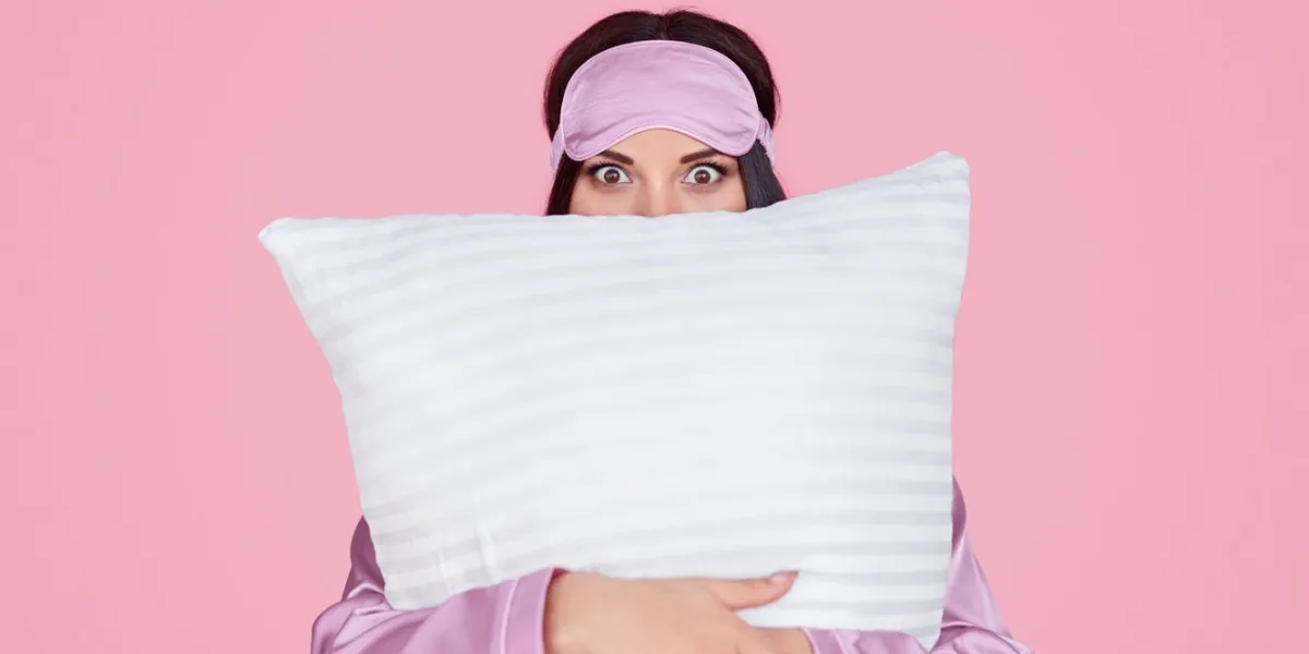 young female in silk lilac sleep mask and pajama looking at camera and embracing comfortable pillow during bedtime against pink background