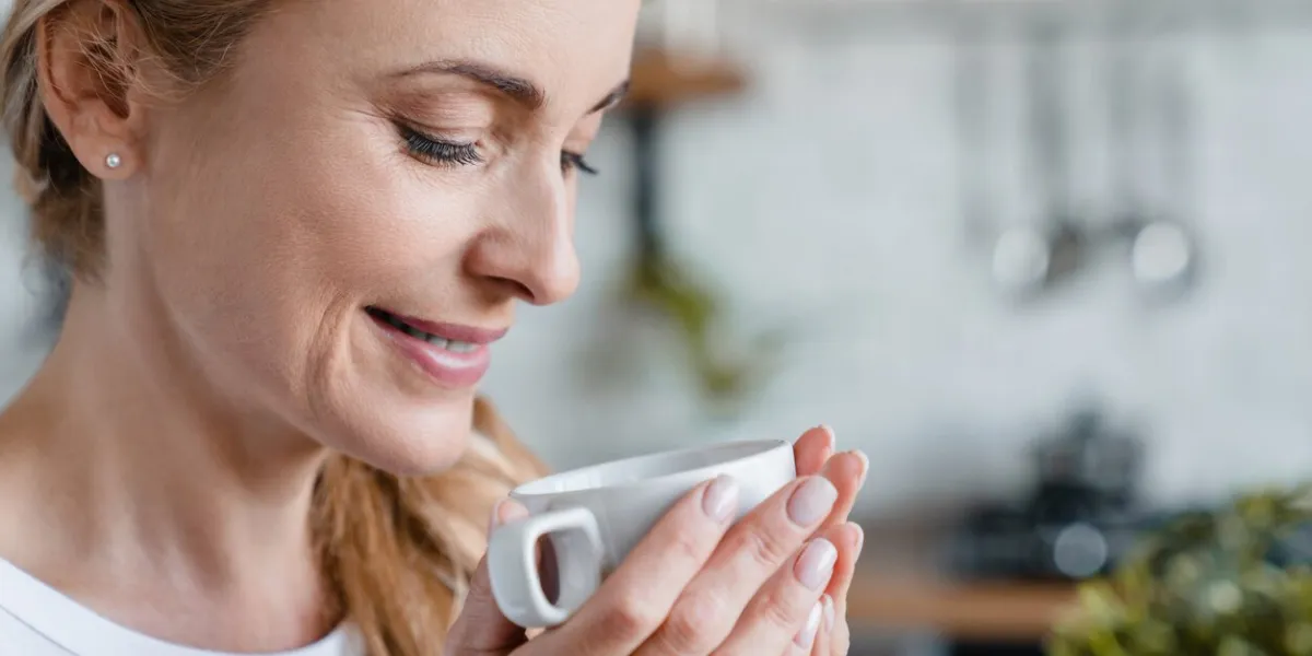 closeup portrait of mature middle-aged woman warming up with coffee tea hot beverage cacao, drinking from a cup at home decaf coffee, feeling smell