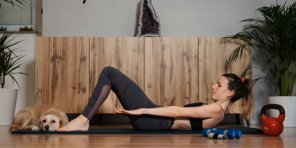 fit young woman lying on an exercise mat in domestic living room doing workout for core strength and belly muscles with her cute pet dog lying at her feet