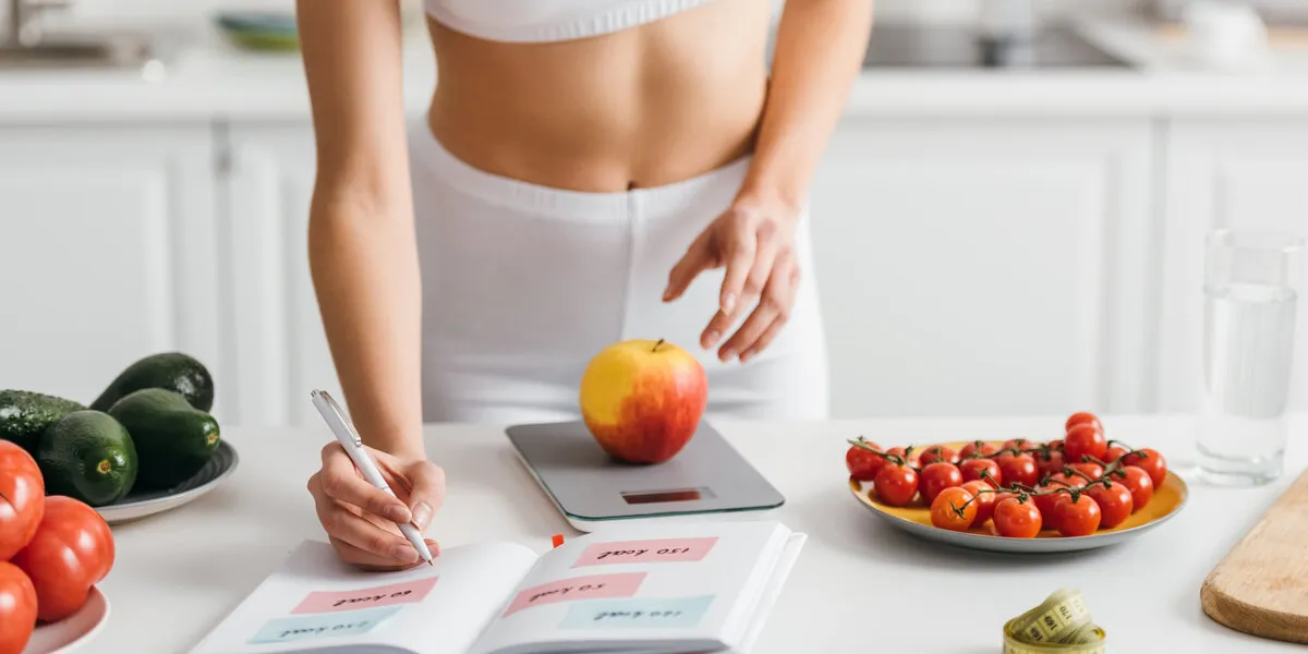 cropped view of fit sportswoman writing calories while weighing apple on kitchen table, calorie counting diet