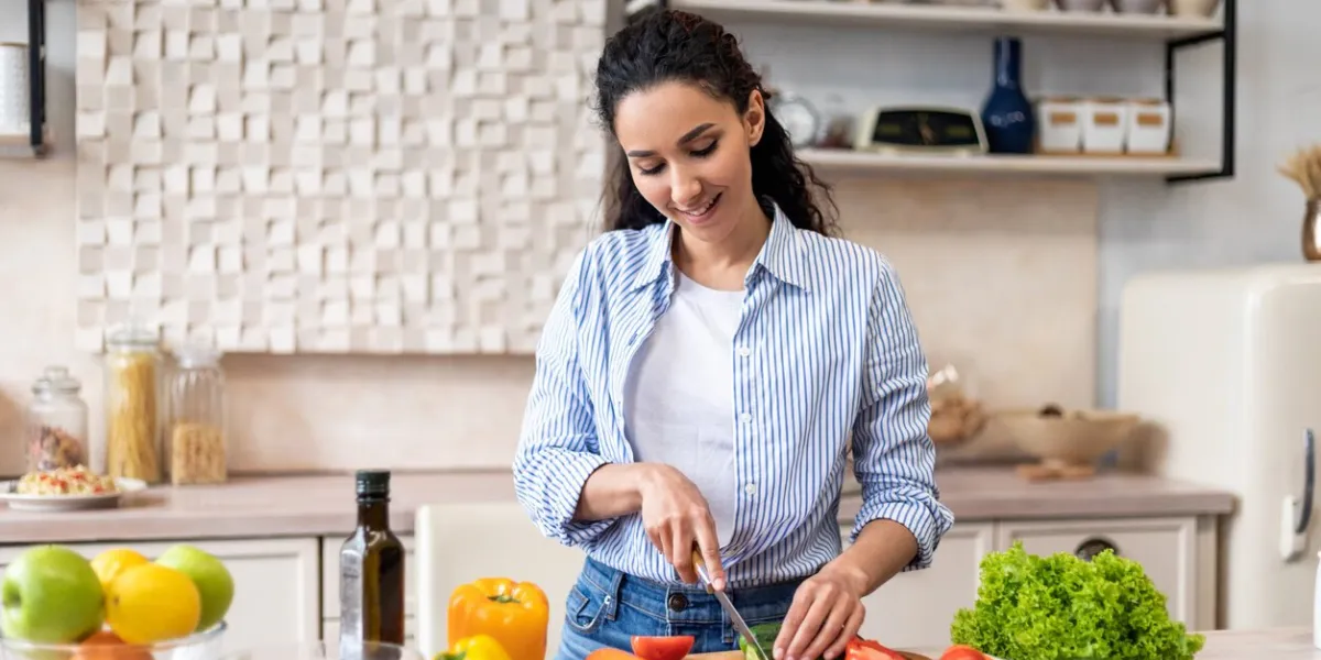 young latin lady cooking delicious and healthy salad standing in modern kitchen, housewife maid preparing vegetable for lunch, slicing cucumber with knife on cutting board