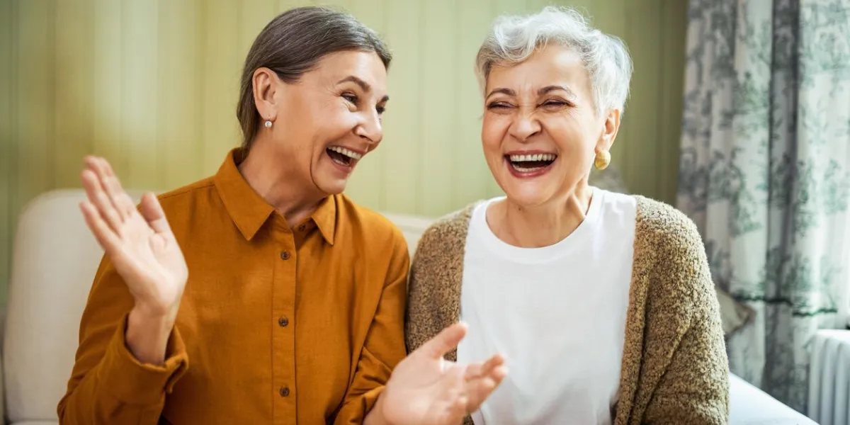 elderly sisters watching comedy together indoors, laughing portrait of two beautiful caucasian women relaxing at home, sitting on sofa, opening mouth wide opened, enjoying weekend, having fun