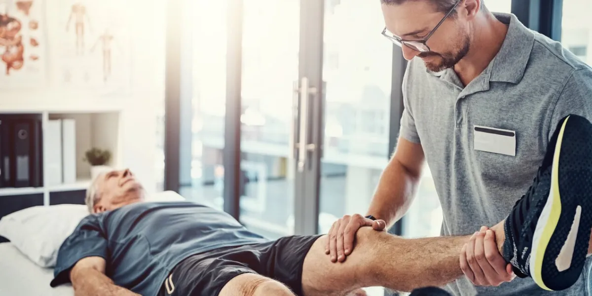 physiotherapy, senior man and stretching leg for knee injury, medical and consultant with healthcare consultation, chiropractor and physical therapy with rehabilitation and care in doctor office