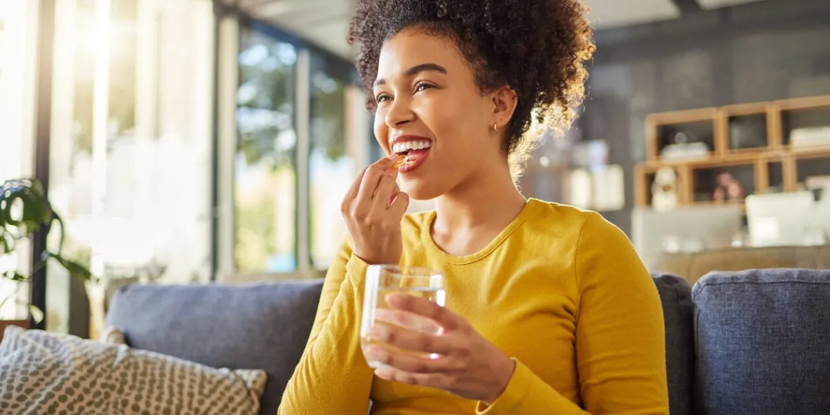 young happy mixed race woman taking medication with water at home one hispanic female with a curly afro taking a vitamin for good health while sitting on the couch at home woman drinking a supplement