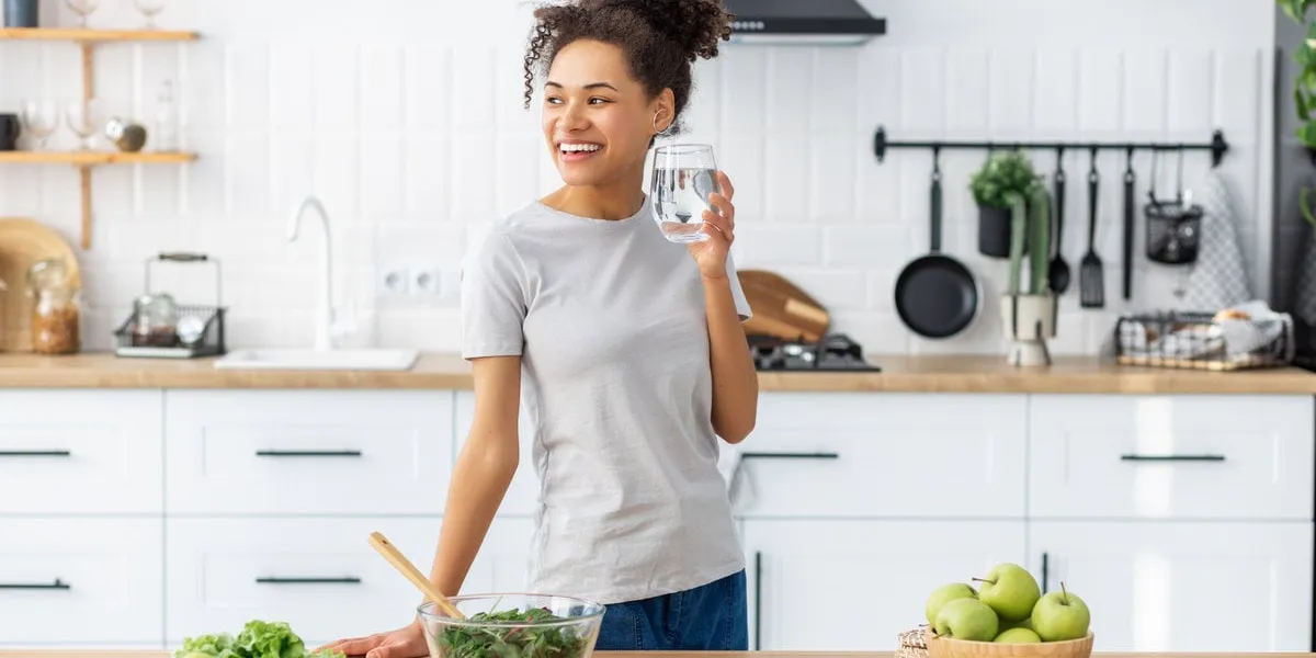 woman cooking healthy food with glass of clean water, looking away and smiling african american young female preparing fresh salad for dinner or breakfast in home kitchen healthy diet eating