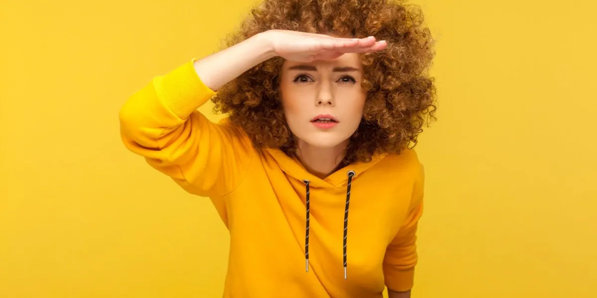 portrait of curious curly-haired woman in urban style hoodie holding hand above eyes and peering into distance, looking far away, expecting and searching someone on horizon studio shot isolated