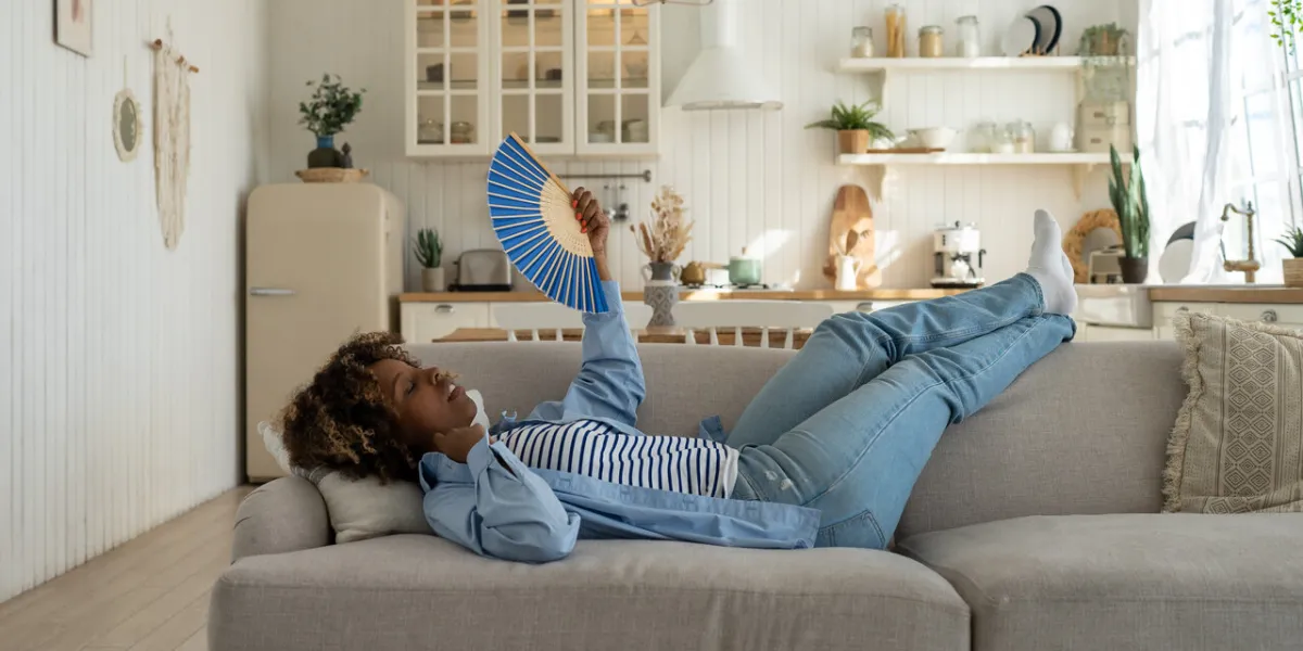unhappy exhausted and overheated young african woman lying on sofa at home waving fan to cool down, black female having hot flashes, feeling hot during heatwave in apartment without air conditioning