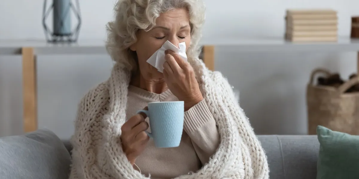 sick mature woman wrapped blanket blowing running nose, feeling unhealthy and ill, upset middle aged female holding paper napkin, handkerchief, holding tea or coffee mug, sitting on couch at home