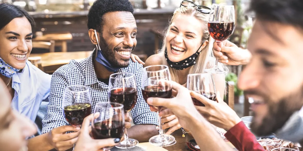 friends toasting red wine at outdoor restaurant bar with open face mask - new normal lifestyle concept with happy people having fun together on warm filter - focus on afroamerican guy
