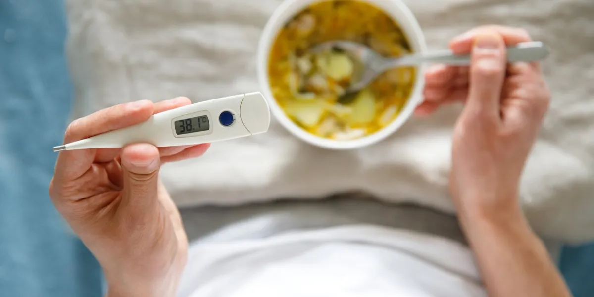 view from above diseased male eating healthy chicken soup to fight the flu sickness, holding digital thermometer in hand, soft focus