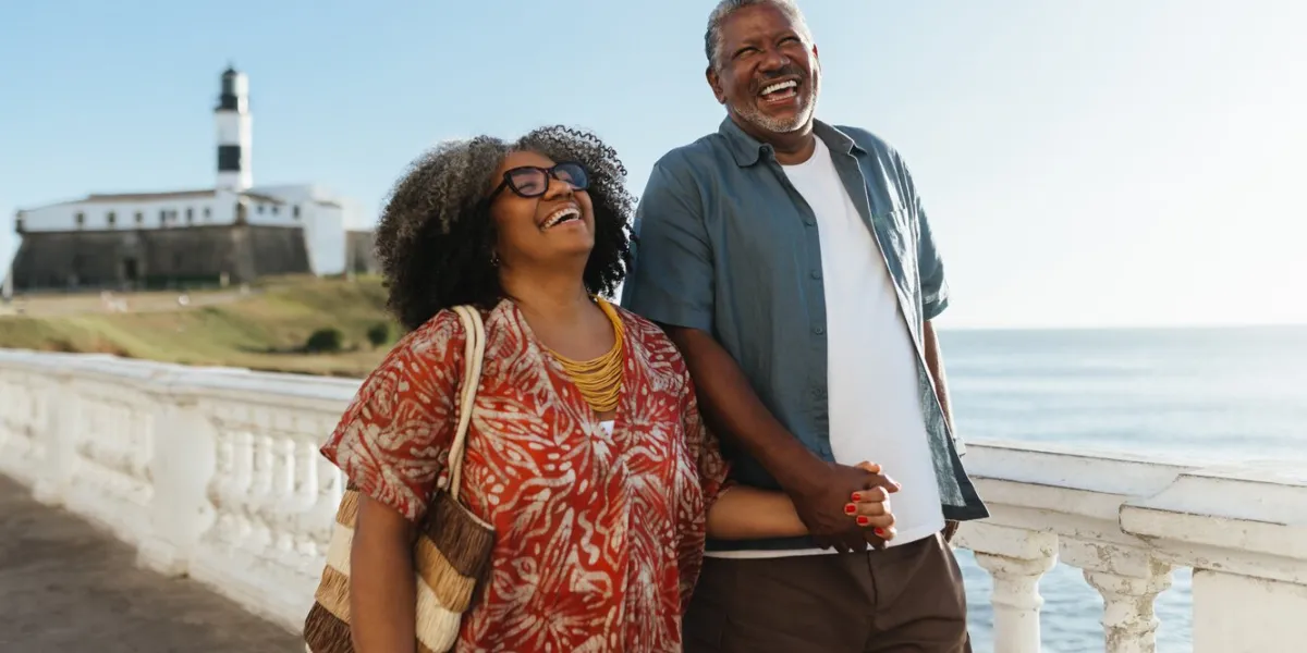 mature pair joyfully holding hands and laughing while enjoying a sunny day by the salvadorian coast with the iconic farol da barra lighthouse in the background