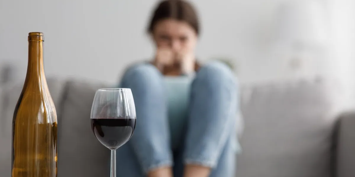 sad caucasian millennial female sits on sofa suffering from stress and troubles, focus on bottle and glass of wine in home interior alcoholism, drinking alone, depressed, addicted to alcohol at home