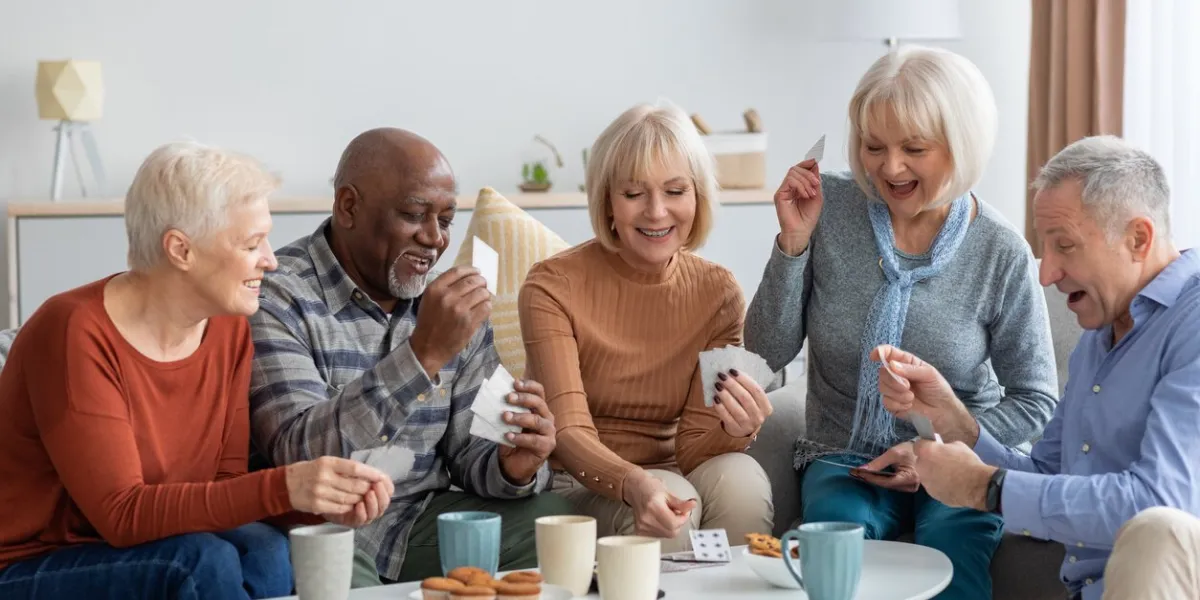 multiethnic group of happy senior friends men and women in casual sitting on couch, having conversation and playing cards while drinking tea with cookies, chilling together at home
