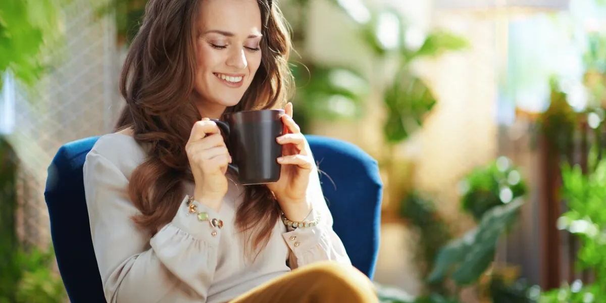 green home happy middle aged woman with long wavy hair with cup of tea sitting in a blue armchair at modern home in sunny day