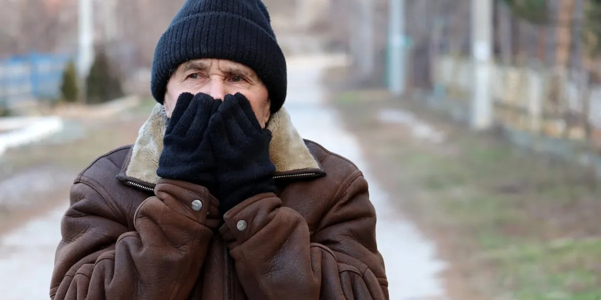 elderly man standing on the rural street and warms up by covering his face with his mittened hands