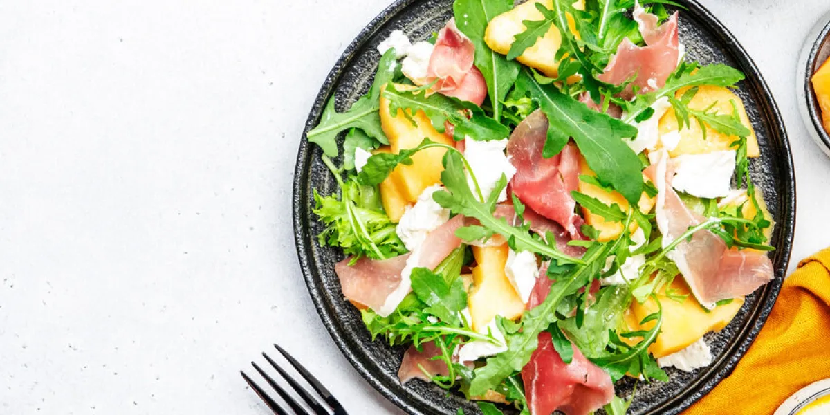 delicious melon salad with cantaloupe, prosciutto, soft cheese and arugula on white table background, top view