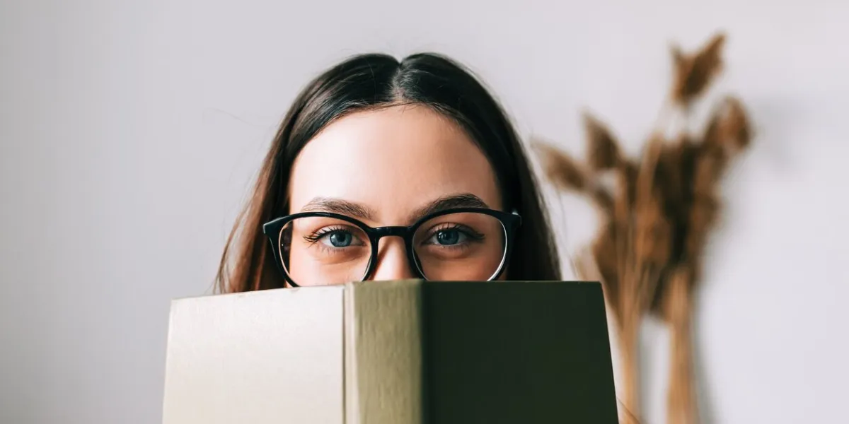 portrait of young caucasian woman college student in eyeglasses hiding behind a book and looking at camera