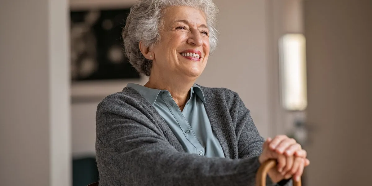 retired senior woman laughing with her wooden walking stick while relaxing at home happy smiling old woman holding walking cane and looking through the window with positivity carefree smiling grandmother sitting on chair and looking away in nursing home