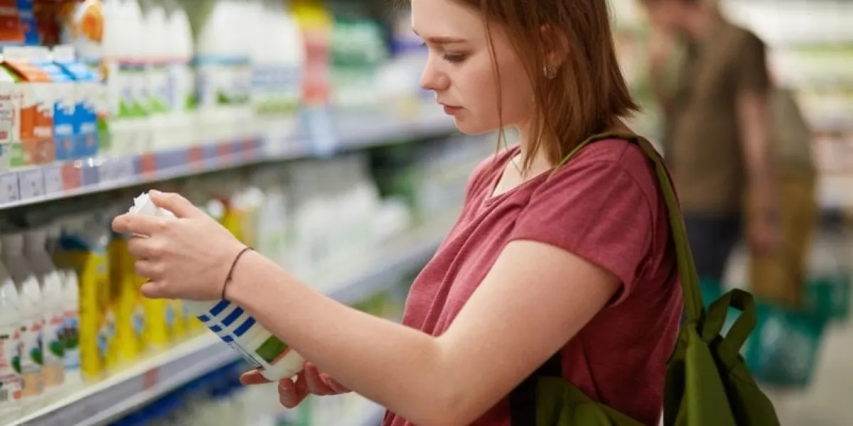 attractive female goes shopping, stands in dairy department, holds bottle of milk, reads information on label, checks date of manufacture, carries rucksack, has serious look, stands in store