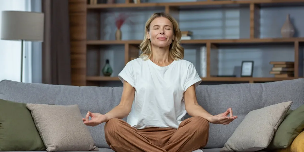 senior woman at home resting meditating with eyes closed sitting on sofa in living room, housewife mature blonde in lotus pose smiling