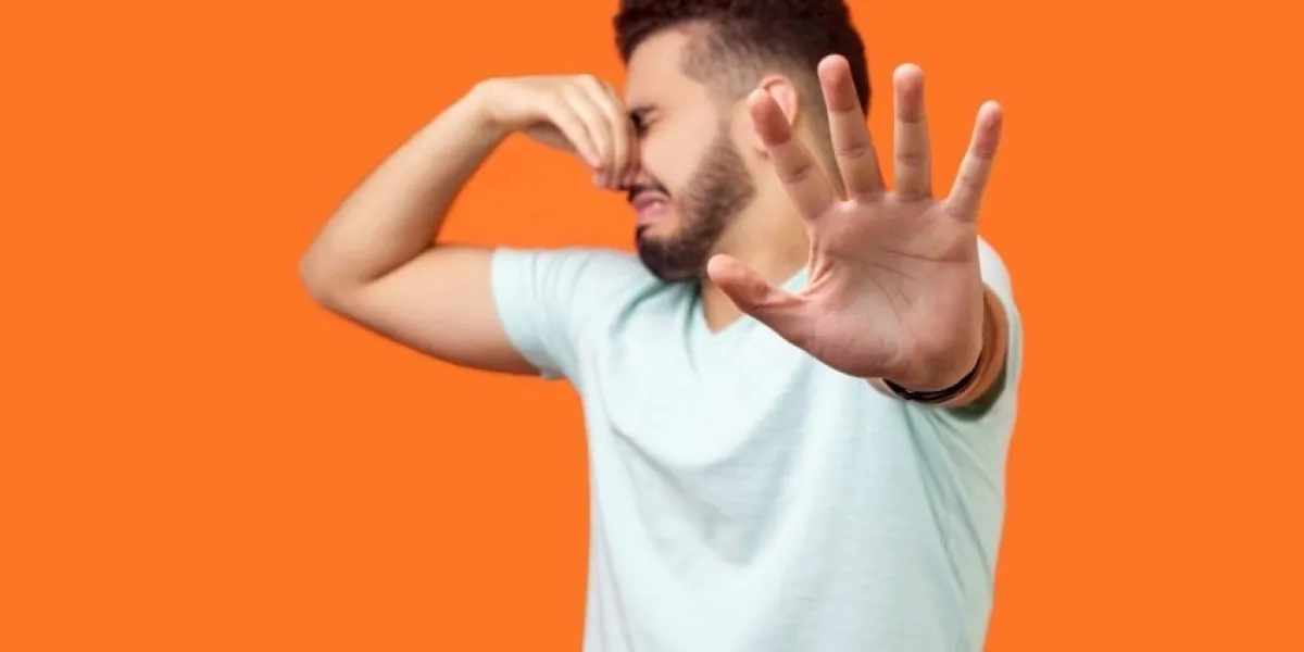 stinky smell side view of confused brunette man with beard in white t-shirt holding breath with fingers on nose, disgusted by bad odor fart, gesturing stop studio shot isolated on orange background