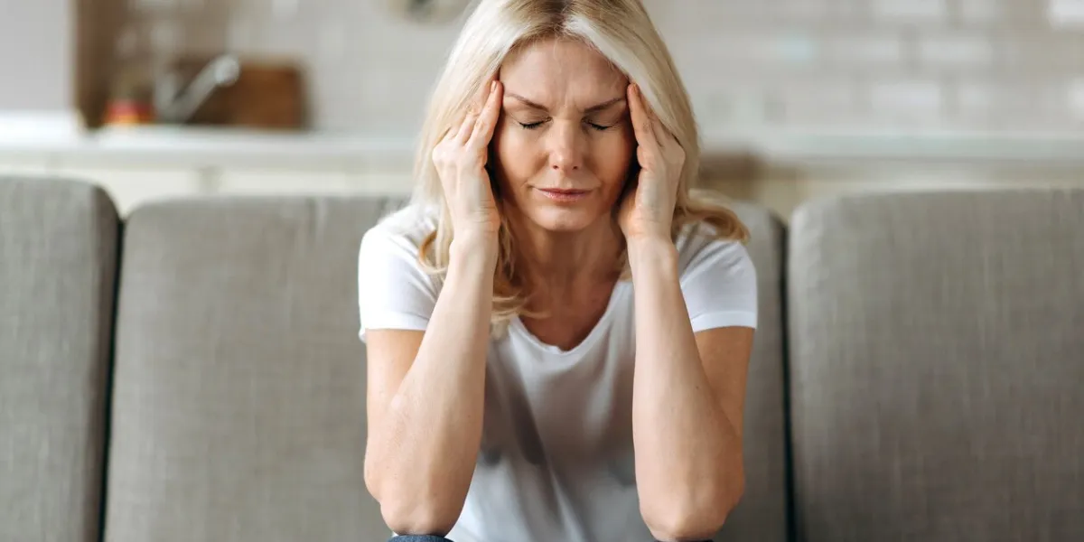 middle aged blonde woman sits on couch at living room holding her head with her hands, feels unhappy because of headache, personal troubles, illness or bad news, she need psychological or medical support