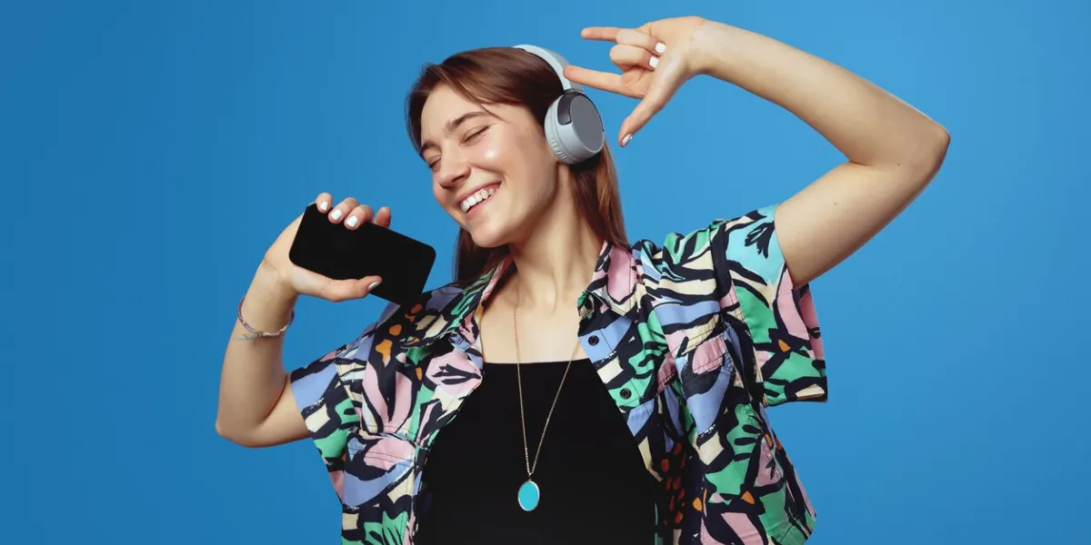 cheerful young girl in headphones smiling and showing rock gesture while holding smartphone with closed eyes, isolated against blue background sign, symbols concept