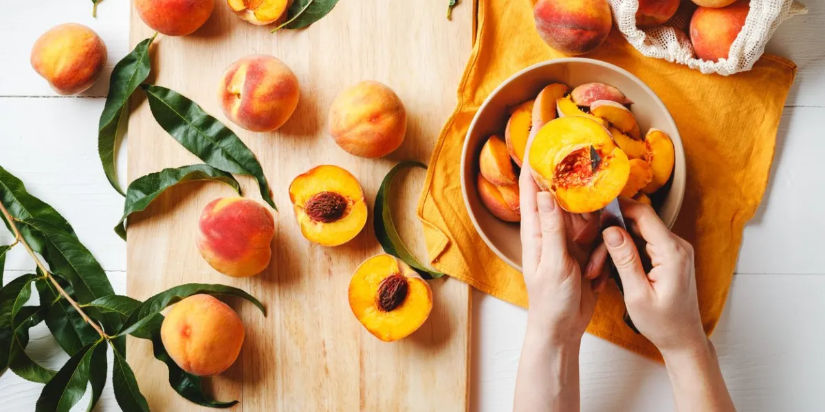 female hands cut fresh delicious ripe peaches into a plate peaches on a cutting board with peach leaves flat lay on a white table with an orange towel